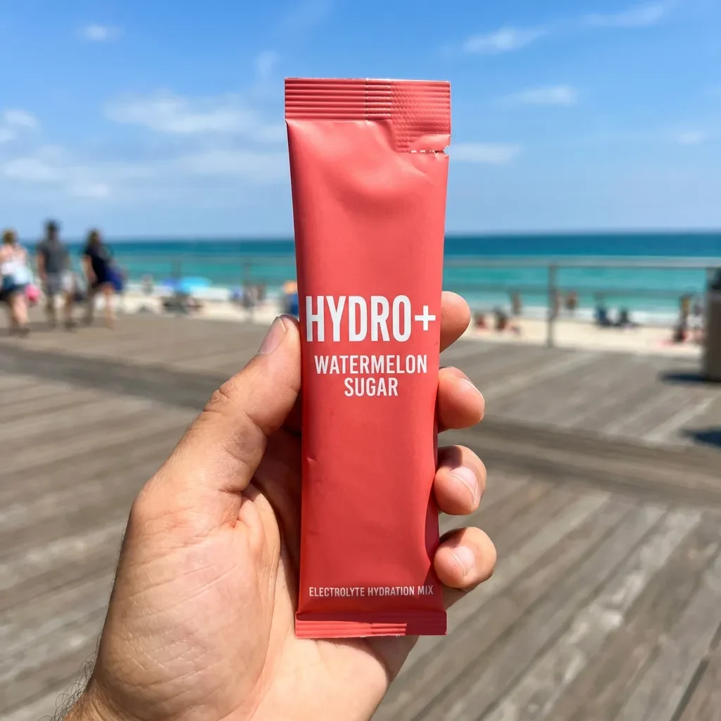 Hand holding a Hydro+ stick pack against a sunlit beach boardwalk